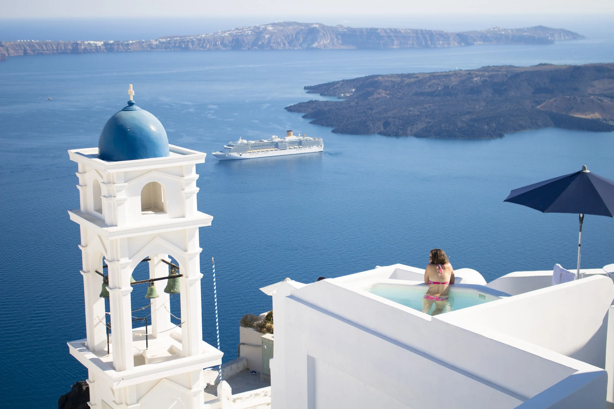 A blue-domed Santorini church above the caldera, with a cruise ship and an infinity pool below